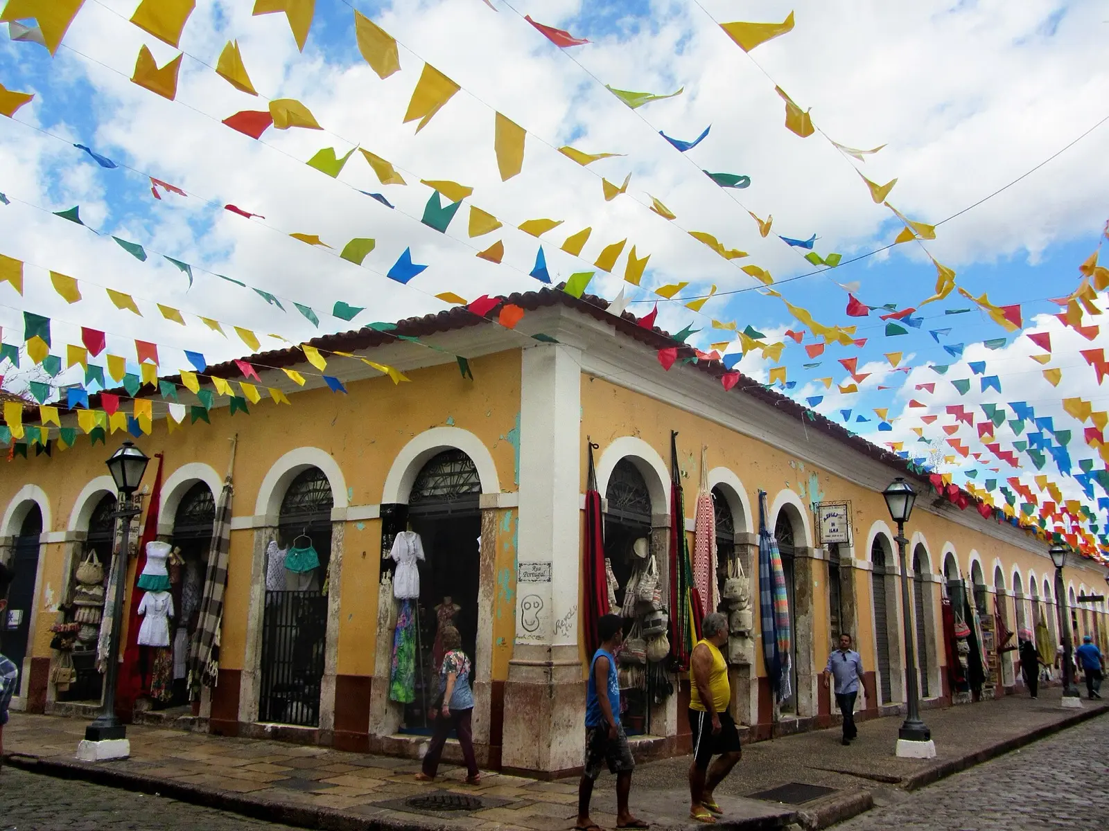 Lojas da Casa das Tulhas, em São Luís, com bandeirinhas coloridas sobre a rua de pedra e pessoas passando.